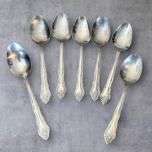 May include: Seven silver spoons with decorative floral patterns on the handles. The spoons have oval bowls and are arranged on a grey surface. The spoons are likely vintage or antique, reflecting a classic design.