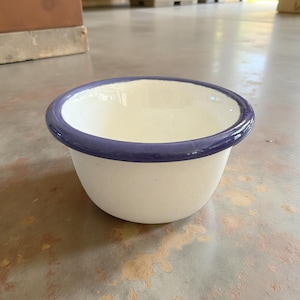May include: A small, white enamel bowl with a rounded shape. The rim is accented with a dark blue band. The bowl is empty and sits on a light-colored surface. This bowl is suitable for serving small portions of food.