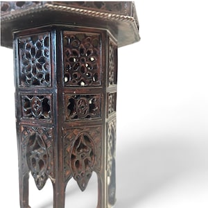 May include: A dark brown, intricately carved wooden table with openwork panels. The table has a hexagonal shape with decorative floral and geometric patterns. The top edge is detailed with a carved border. The legs are arched.