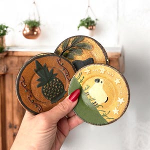May include: Set of three decorative coasters with painted designs. One features a pineapple, another has a leaf design, and the third has a white figure with stars. The coasters are brown, green, and yellow.