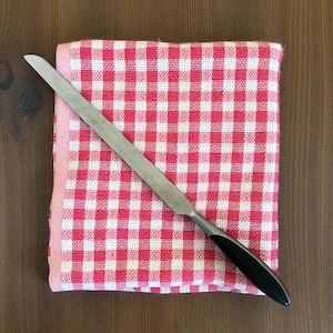 May include: A stainless steel cake knife with a black handle rests on a red and white checkered kitchen towel. The towel is folded and placed on a wooden surface. The knife is angled across the towel.