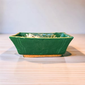 May include: A rectangular green ceramic bowl with a flared rim and a textured floral relief pattern on the exterior sides. The base of the bowl is a contrasting tan color, and the interior features a glossy finish with visible light reflections.