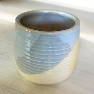 May include: A handmade ceramic tumbler in light blue and cream. The exterior has horizontal ridges, adding texture. The rim and interior are a darker blue, contrasting with the lighter body. A unique, handcrafted piece.