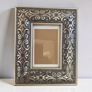 May include: A rectangular picture frame featuring a metallic silver and dark grey finish. The wide border displays an intricate embossed pattern of swirling scrolls and floral motifs set against a textured, stippled background.