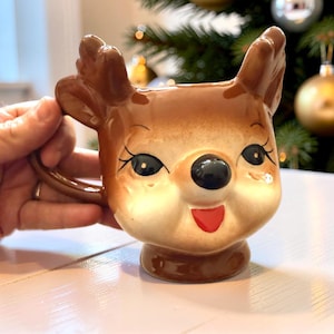 May include: A ceramic mug shaped like a reindeer head with brown fur, large dark eyes with long eyelashes, a black nose, and a small red mouth. The mug features two brown antlers extending from the top and a handle on the side for holding.