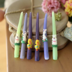 May include: Six Easter-themed taper candles in shades of green, purple, and violet. Each candle features a decorative base with a bunny or chick figurine. The candles are arranged on a wooden surface, with flowers in the background.
