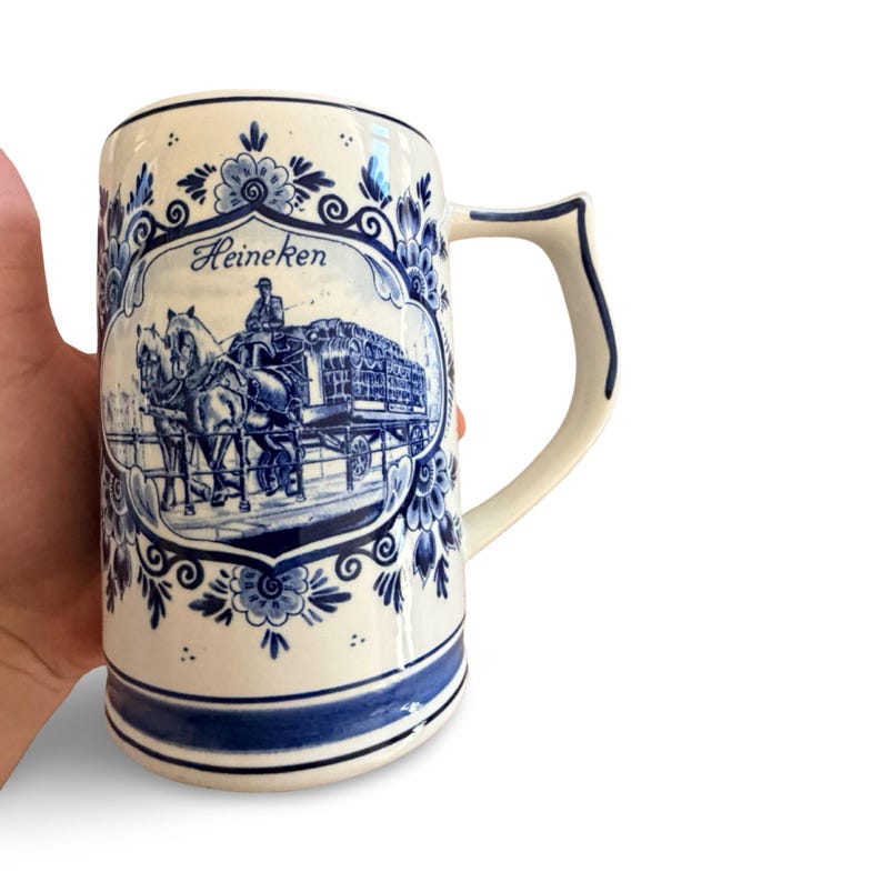 May include: A white ceramic beer stein with blue floral and scenic designs. The stein features a blue and white illustration of a horse-drawn carriage with the word "Heineken" in a blue oval. The handle is white with blue trim.