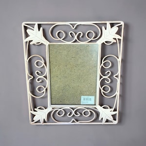 May include: A rectangular picture frame with a distressed off-white finish, featuring a metal border with scrollwork patterns and leaf-shaped corner accents. The centre contains a plain backing board with a small rectangular product label.
