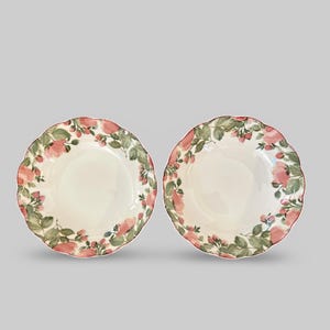 Puede incluir: Dos platos de cerámica blanca con borde festoneado. Cada plato está decorado con un motivo floral de rosas rosas y hojas verdes alrededor del borde. Los platos tienen un borde rojo.