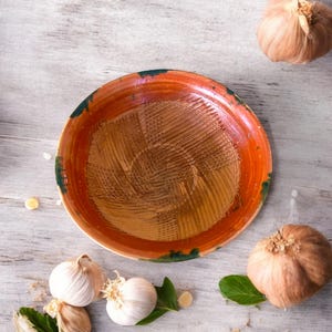 May include: A handmade, rustic ceramic bowl with a textured interior and a vibrant orange and green rim. The bowl is surrounded by fresh garlic bulbs and green leaves, suggesting a culinary theme.