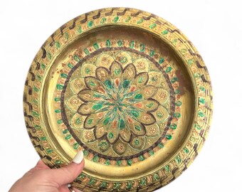 May include: An antique brass tray with a detailed floral pattern. The tray has green, red, and brown accents, and a decorative border. The tray is held by a hand. The tray's diameter is approximately 25 cm.