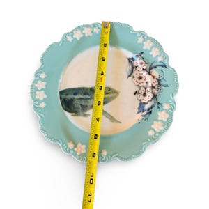 Pu&ograve; includere: Un piatto rotondo in ceramica azzurro con bordo smerlato e decorazioni floreali bianche in rilievo. Al centro sono dipinti un camaleonte verde e dei fiori bianchi. Un metro a nastro giallo indica un diametro di 28 cm.