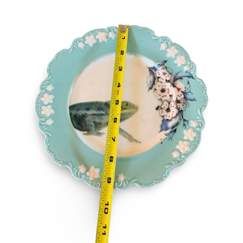 Pu&ograve; includere: Un piatto rotondo in ceramica azzurro con bordo smerlato e decorazioni floreali bianche in rilievo. Al centro sono dipinti un camaleonte verde e dei fiori bianchi. Un metro a nastro giallo indica un diametro di 28 cm.