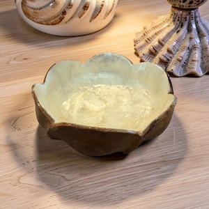 May include: A decorative, handcrafted ceramic bowl with a unique, scalloped edge. The bowl features a light yellow interior and a dark brown exterior. The bowl is sitting on a wooden surface. Other ceramic pieces are in the background.