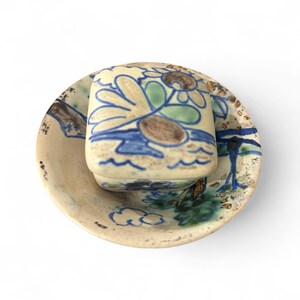 May include: A ceramic teacup and saucer set. The cup and saucer are painted with blue, green, and brown floral and abstract designs on a cream background. The set has a rustic, handmade appearance.