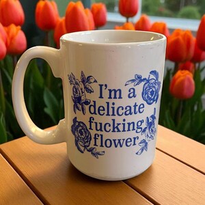 以下が含まれることがあります： 青い花の模様と「I'm a delicate fucking flower」の文字が入った白いセラミックマグ。マグには大きなハンドルが付いています。背景にはオレンジ色のチューリップがあります。