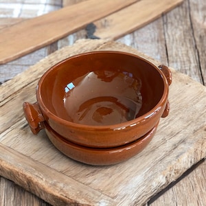 May include: A pair of brown ceramic bowls with handles. The bowls are round and have a glossy finish. They are stacked on top of each other. The bowls are sitting on a wooden surface.