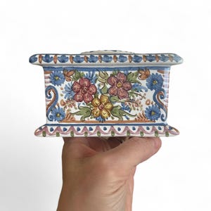 Könnte beinhalten: Eine rechteckige Keramikbox mit floralem Design. Die Box hat eine weiße Basis mit blauem Rand und ist mit roten, gelben und blauen Blumen verziert. Die Box wird von einer Hand vor weißem Hintergrund gehalten.