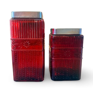 Op de afbeelding: Twee rode glazen potten met zilverkleurige deksels. De grotere pot heeft verticale lijnen en een decoratief bloemmotief. De kleinere pot heeft een vergelijkbaar ontwerp. Beide zijn rechthoekig.