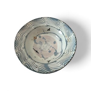 May include: A ceramic bowl with a wavy, blue and white rim design. The interior features a light pink and blue abstract pattern with small black bird-like shapes. The bowl has a glossy finish and is set against a white background.