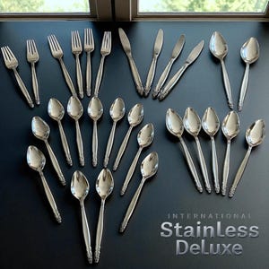 May include: A collection of stainless steel cutlery, comprising forks, knives, and spoons, displayed on a dark surface. The silverware features a decorative handle pattern. The text "INTERNATIONAL Stainless Deluxe" is present.
