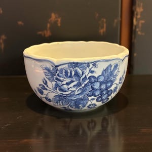 Puede incluir: Un cuenco de cerámica blanca con borde festoneado y diseño floral azul. El cuenco presenta un patrón detallado de rosas y uvas, creando una estética clásica y elegante. El interior del cuenco es de color blanco crema.