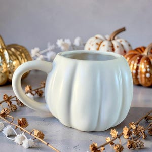 May include: A white ceramic mug shaped like a pumpkin with a curved handle. The mug has vertical ridges and sits on a surface with decorative fall elements, including other pumpkin-shaped decorations in gold and orange.