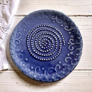 May include: A round, blue ceramic plate with a spiral design in the center. The plate has a textured surface and decorative swirls around the edge. The plate is on a white wooden surface.