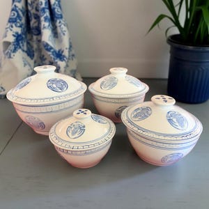 May include: Four white ceramic bowls with lids, featuring blue decorative patterns and bird motifs. The bowls vary in size, with a matching design on the lids and rims. A blue and white patterned cloth is visible in the background.