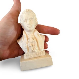 May include: A small, off-white bust of Wolfgang Amadeus Mozart, featuring a detailed sculpted face, period-style cravat, and collared coat. The base of the sculpture is rectangular and clearly displays the embossed text MOZ ART on the front.