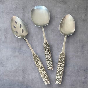 Può includere: Tre cucchiai da portata d'argento con manici incisi. Un cucchiaio ha una coppetta forata, un altro è un cucchiaio da portata pieno e il terzo ha una coppetta arrotondata. I cucchiai sono su una superficie grigia.