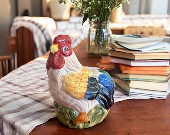 Pode incluir: Uma estatueta de galo de cerâmica com crista e barbela vermelhas, bico amarelo e plumagem colorida. O galo está pousado sobre uma base verde. A estatueta está sobre uma mesa de madeira com uma toalha de mesa xadrez.