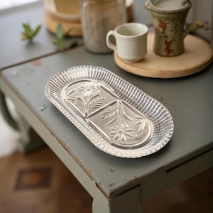 May include: Clear glass oval serving tray with a floral design. The tray has a ribbed border and is decorated with etched flowers and leaves. The tray is on a gray table.