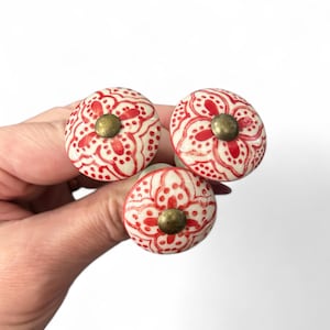 Op de afbeelding: Drie decoratieve kastknoppen. Elke knop heeft een witte basis met een rood bloemmotief en een messingkleurig midden. De knoppen zijn rond en hebben een vintage of bohemien stijl.