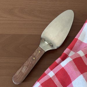 May include: A stainless steel pie server with a wooden handle. The handle is attached with two brass rivets. A red and white checkered cloth is in the background. The server is on a wooden surface.