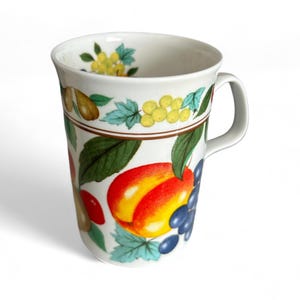 May include: White ceramic mug with a colorful fruit design. The mug features illustrations of peaches, grapes, pears, and other fruits, with green leaves and yellow accents. A brown band separates the fruit design from the rim.