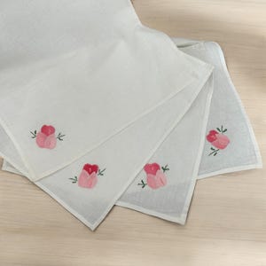Puede incluir: Pañuelos de lino blanco con rosas rosas y hojas verdes bordadas. Los pañuelos están doblados y dispuestos en forma de abanico, mostrando el bordado floral en cada esquina. El fondo es una superficie de madera clara.