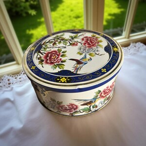 Può includere: Scatola rotonda vintage con un design floreale e di uccelli. Il coperchio presenta fiori rossi e bianchi, un uccello blu e un bordo blu. I lati della scatola presentano anche motivi floreali e di uccelli. La scatola è bianca con dettagli dorati.