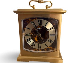 May include: A gold-toned Howard Miller quartz clock with a rectangular shape. The clock face features Roman numerals and a dark brown marble-like background. A gold handle is on top. The clock is labeled "Japan".