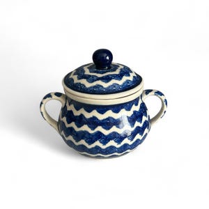 May include: A ceramic sugar bowl with a lid, featuring a blue and white wave pattern. The bowl has two handles and a small knob on the lid. The design is repeated on the lid and the body of the bowl.