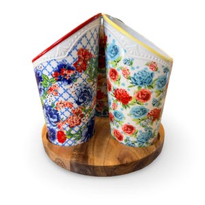 May include: A set of three ceramic utensil holders with floral patterns in red, blue, and white. The holders sit on a round wooden base. The rims of the holders are trimmed in red and yellow.