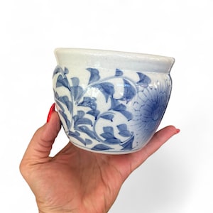 以下が含まれることがあります： 青い花の模様が施された、小さな白い陶器の植木鉢。植木鉢には、青い花と葉の模様があります。縁は明るい白です。植木鉢は手で持たれています。