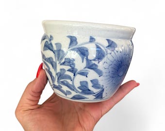May include: A small, white ceramic planter with blue floral designs. The planter features a blue flower and leaf pattern. The rim is a lighter shade of white. The planter is held in a hand.