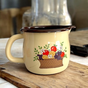 Op de afbeelding: Crèmekleurige emaille kan met een bruine rand en handvat. De voorkant heeft een illustratie van een fruitmand, met rode appels, paarse druiven en witte bloemen. De kan staat op een houten oppervlak.