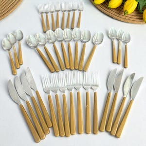May include: A collection of stainless steel silverware with light brown wooden handles. The set includes forks, spoons, and knives, arranged on a white surface. The silverware is designed for dining and serving.