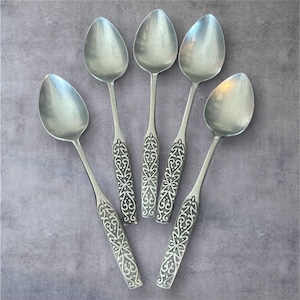 May include: Set of five silver spoons with ornate, engraved handles. The spoons have a classic design with oval bowls and detailed floral patterns on the handles. The spoons are arranged on a gray surface.