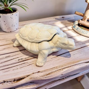 Puede incluir: Un joyero de cerámica color crema con forma de tortuga y tapa extraíble. La tortuga presenta un patrón de caparazón segmentado y texturizado con un acabado liso y brillante. La pieza está esculpida con cuatro patas y una cabeza extendida.