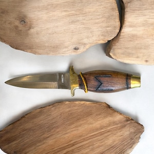 May include: A dagger with a brass blade and handle, featuring a wooden grip with a dark etched design. The handle is accented with brass fittings and a yellow cord. The blade has the word "Pakistan" etched on it.