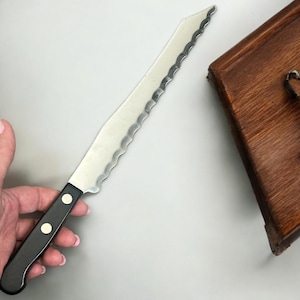 May include: A serrated bread knife with a black handle and two silver rivets. The stainless steel blade has a wavy edge. A wooden cutting board is in the background.
