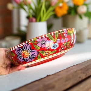 Pode incluir: Uma tigela de cerâmica vermelha com um design floral. A tigela apresenta um padrão vibrante de flores coloridas, incluindo roxas, rosas, amarelas e azuis, sobre um fundo vermelho. A borda tem um padrão listrado.